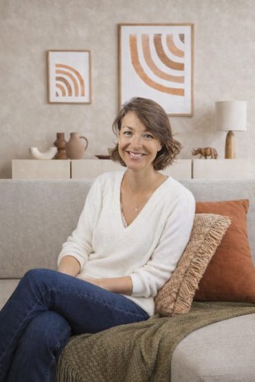 Sandra Devaux, décoratrice d'intérieur sur Nice, pose dans son canapé dans son domicile avec une jolie décoration couleurs crème, beige et terra cotta en fond.